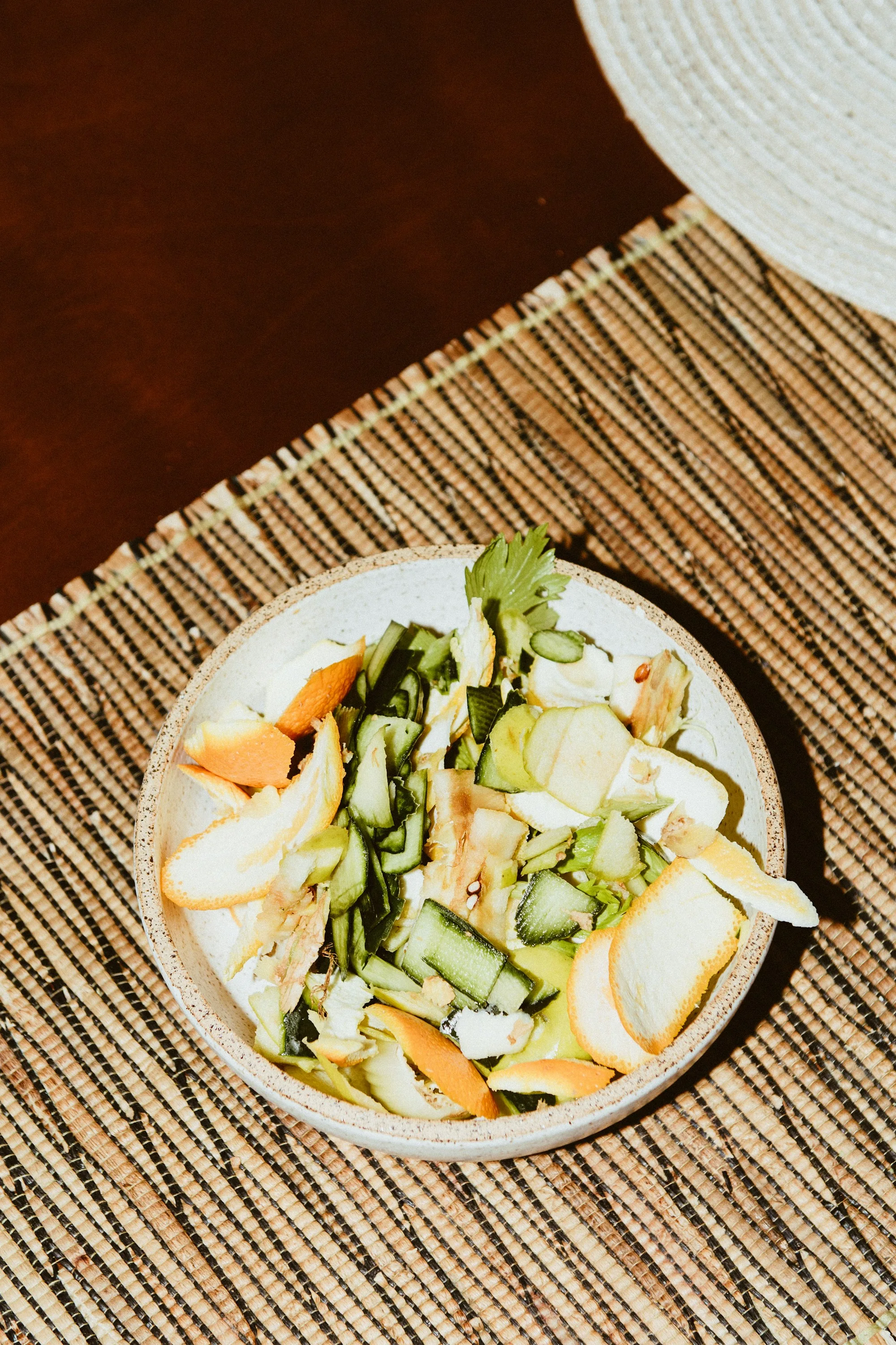 A small ceramic bowl of vegetable trim and herb stems on a woven mat — the food waste hiding in every restaurant's prep